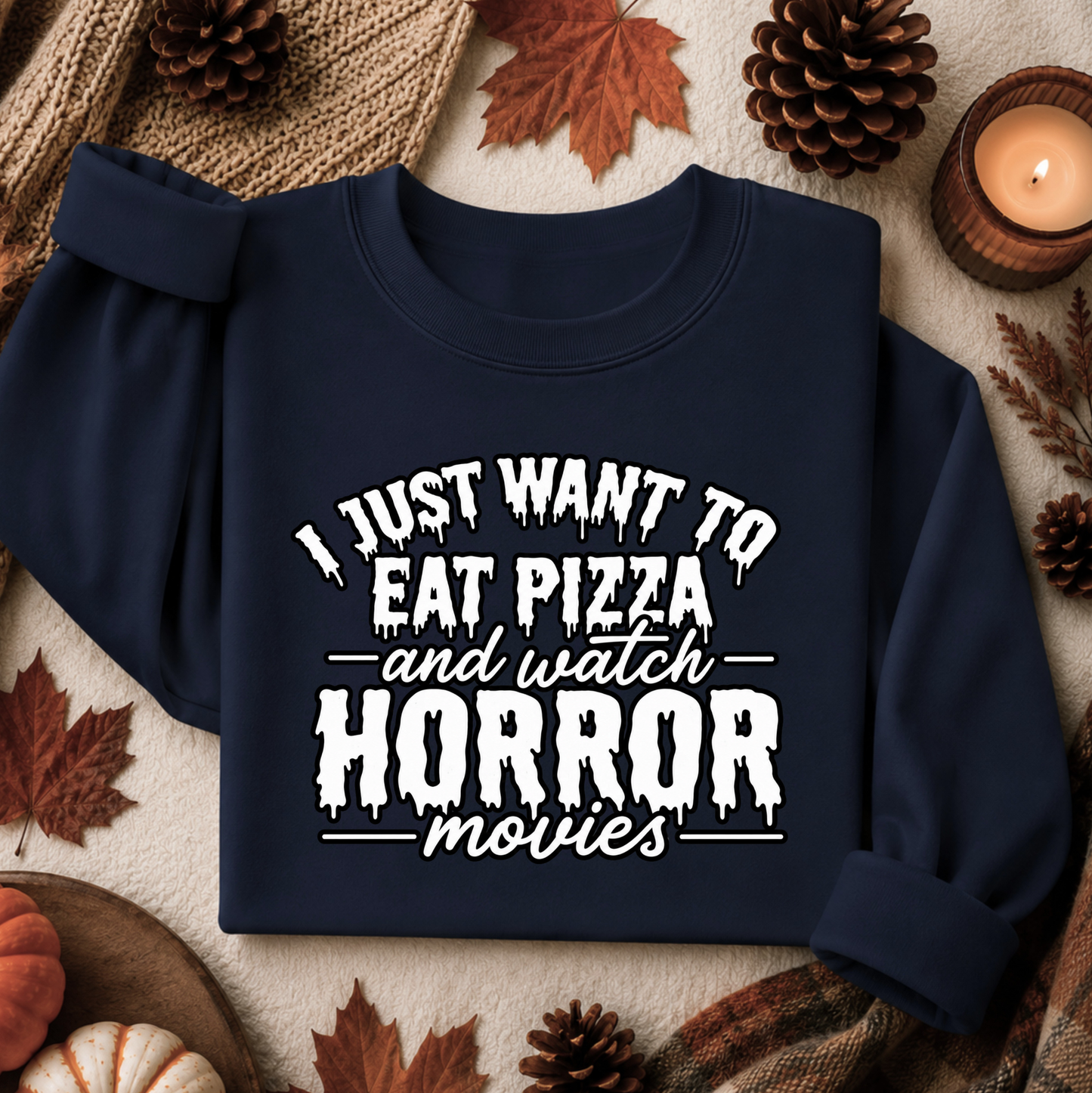 A navy blue sweatshirt with a white graphic text reading 'I JUST WANT TO EAT PIZZA and watch HORROR movies' laid flat on a cozy autumn-themed background with pinecones, leaves, and a candle.