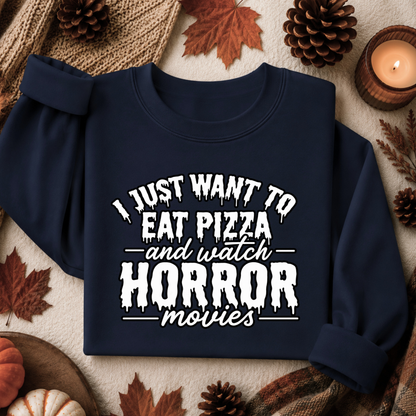 A navy blue sweatshirt with a white graphic text reading 'I JUST WANT TO EAT PIZZA and watch HORROR movies' laid flat on a cozy autumn-themed background with pinecones, leaves, and a candle.