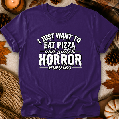 A purple short-sleeve graphic T-shirt laid flat with a white horror-movie pizza slogan, surrounded by autumn props like pinecones, leaves, a candle, and a small pumpkin.