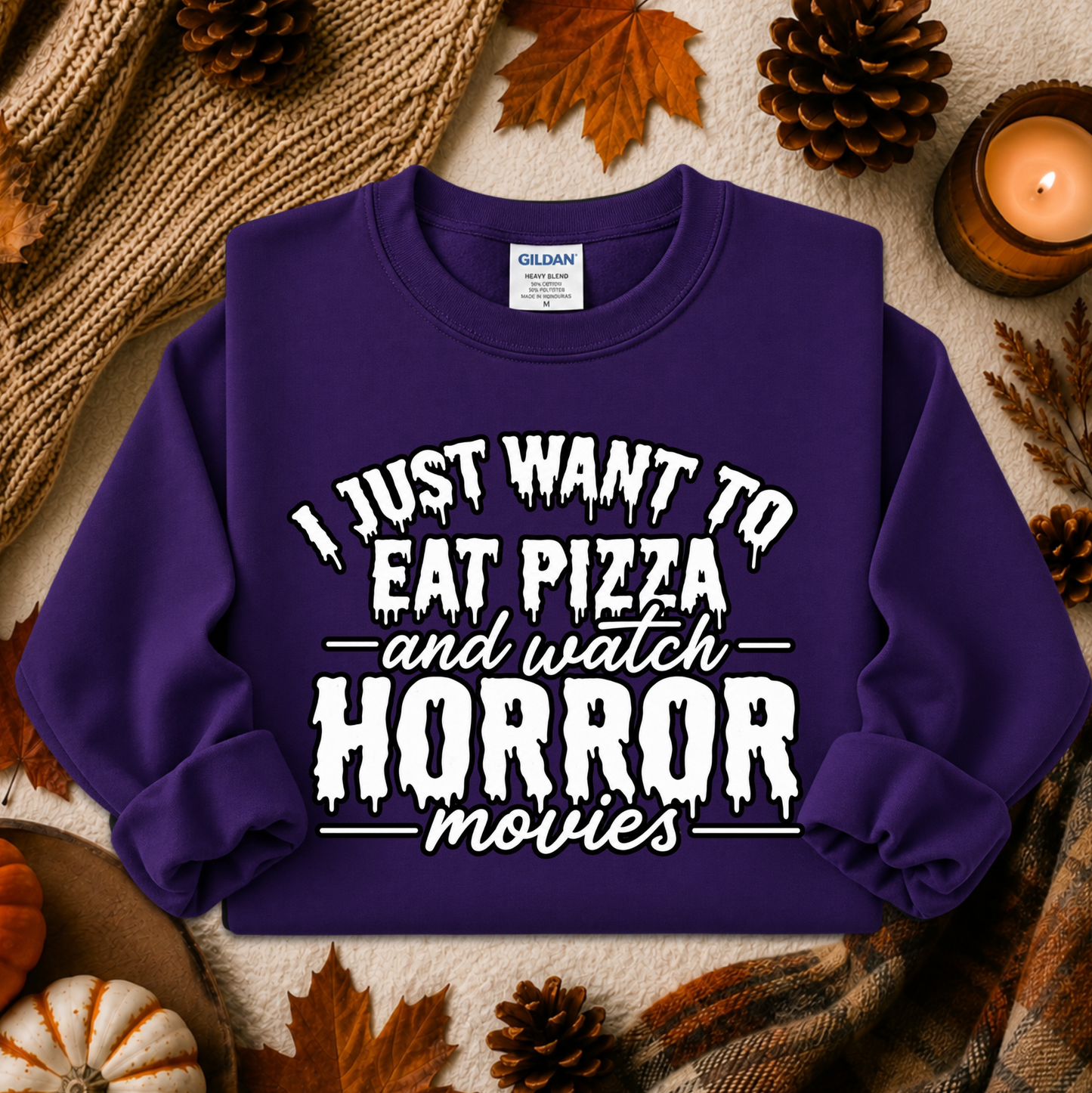 A purple long-sleeve sweatshirt with a white dripping text graphic reading 'I JUST WANT TO EAT PIZZA and watch HORROR movies' laid flat on an autumn-themed background with leaves, pinecones, a candle, and a knit blanket.