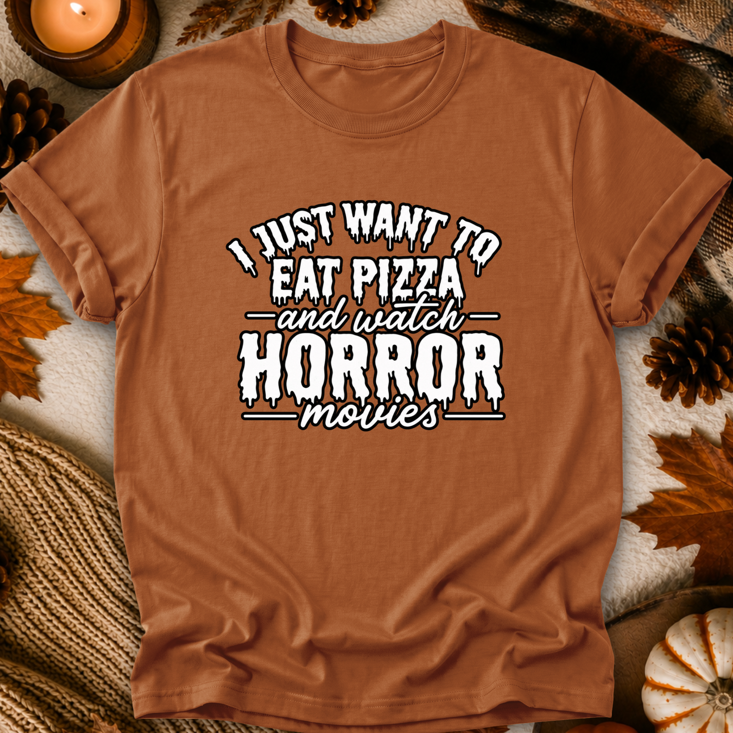 A rust-brown short-sleeve t-shirt with a white graphic text reading 'I JUST WANT TO EAT PIZZA and watch HORROR movies', styled flat-lay with autumn props like pinecones, leaves, a candle, and a knit blanket.