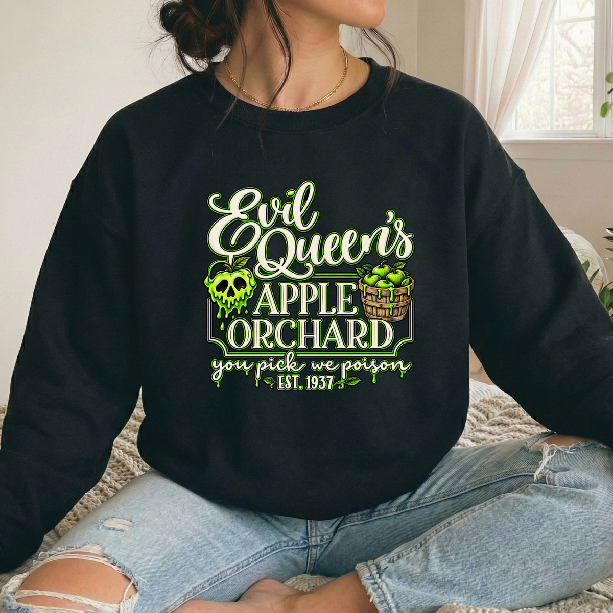 A person wearing a black crewneck sweatshirt with a green and white graphic that reads 'Evil Queen's Apple Orchard you pick we poison Est. 1957', paired with ripped light-wash jeans in a softly lit indoor setting.