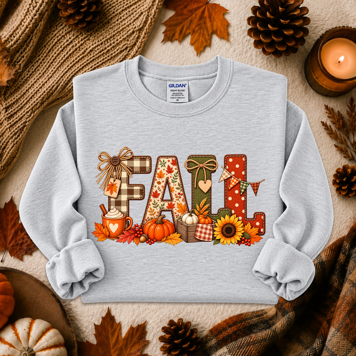 A light gray crewneck sweatshirt with a colorful 'FALL' graphic print, styled in a flat lay with autumn props including pinecones, leaves, a candle, and a knitted scarf.