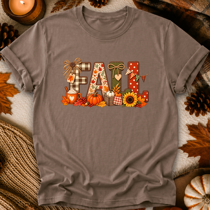 A taupe gray short-sleeve t-shirt with a colorful 'FALL' graphic print, styled in a flat-lay with autumn props like pine cones, a lit candle, and leaves.