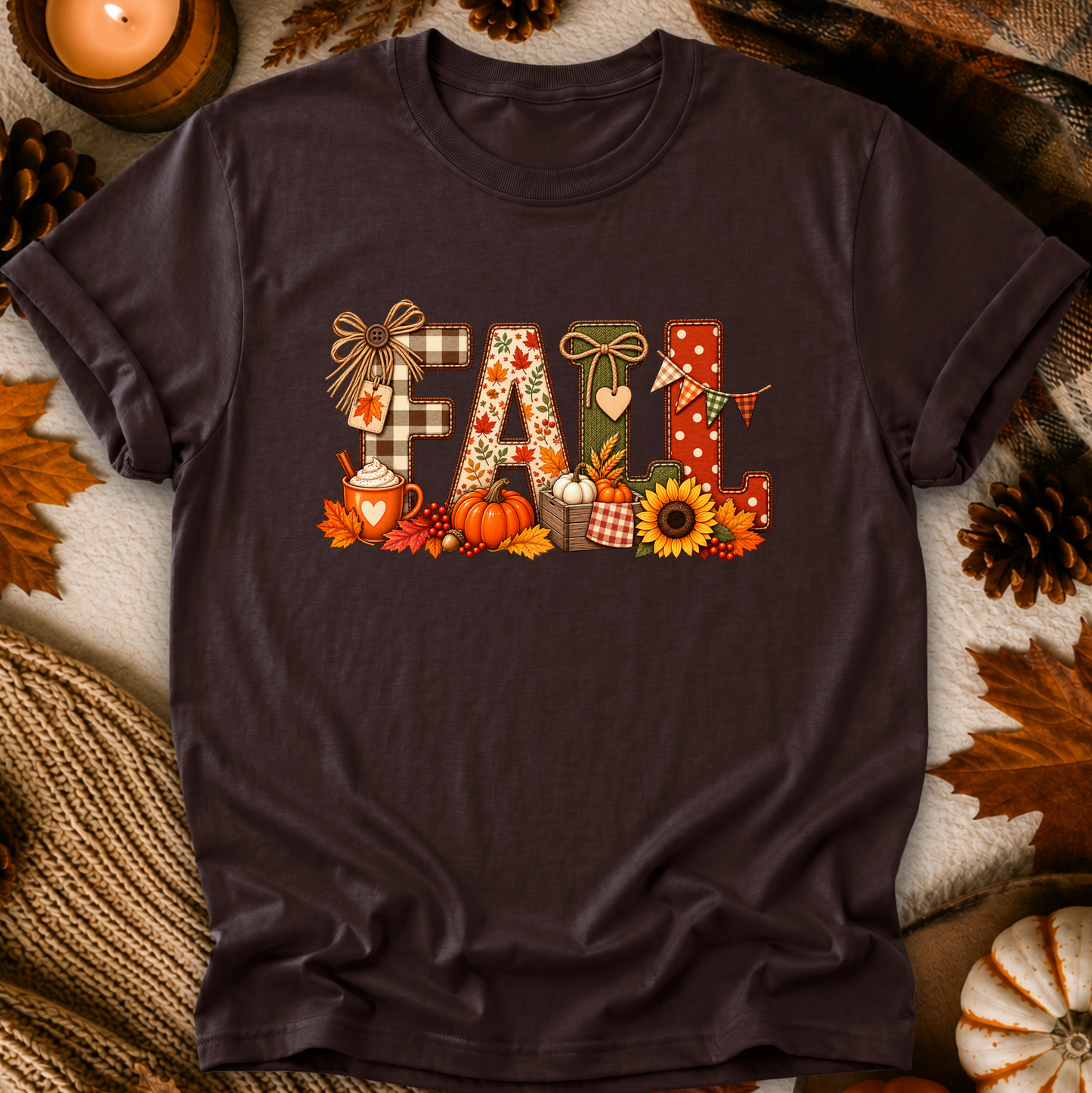 A dark brown short-sleeve t-shirt with a colorful 'Fall' graphic print, styled in a flat-lay arrangement with autumn leaves, pinecones, a candle, and a knitted blanket.
