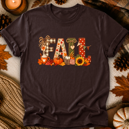A dark brown short-sleeve t-shirt with a colorful 'Fall' graphic print, styled in a flat-lay arrangement with autumn leaves, pinecones, a candle, and a knitted blanket.