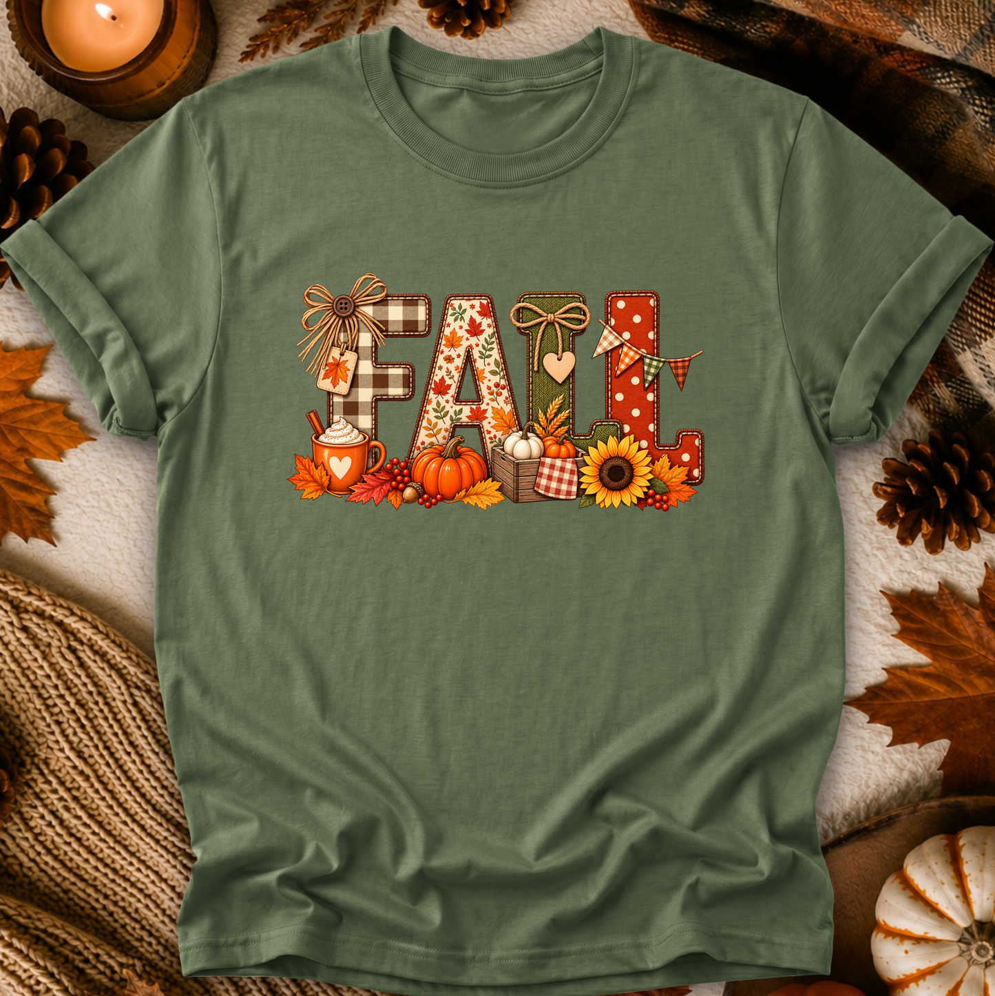 An olive green short-sleeve t-shirt with a 'Fall' graphic print, styled in a flat-lay arrangement with autumn leaves, pinecones, a candle, and a cozy knit blanket.