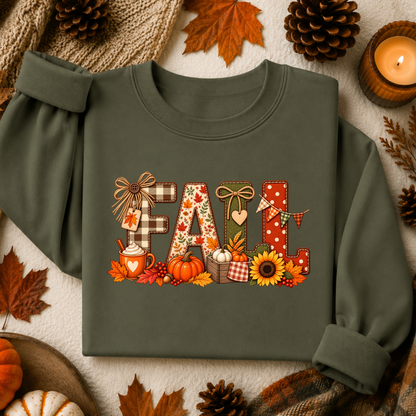 An olive green long-sleeve sweatshirt with a festive 'FALL' graphic print, styled in a flat-lay arrangement with autumn leaves, pinecones, a candle, and pumpkins on a textured cream background.