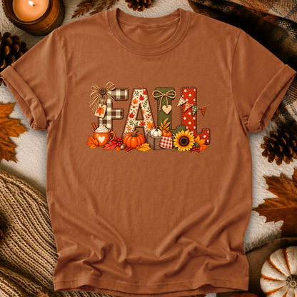 A rust-brown short-sleeve t-shirt laid flat with a decorative 'FALL' graphic made from plaid, polka dots, leaves, pumpkins, sunflowers, and autumn motifs. The shirt is styled with fall props including pinecones, leaves, a candle, and a knit blanket.