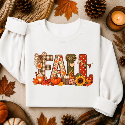 A white long-sleeve sweatshirt laid flat with rolled cuffs, featuring a colorful autumn-themed 'FALL' graphic on the chest. The design includes plaid, polka dots, leaves, pumpkins, sunflowers, and a mug, surrounded by fall leaves, pinecones, a candle, and a knit scarf.