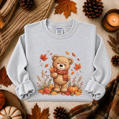 A light gray long-sleeve crewneck sweatshirt with a graphic of a cute cartoon teddy bear wearing a red scarf, surrounded by autumn leaves, pumpkins, and fall foliage. The sweatshirt is styled in a cozy flat-lay arrangement with a knitted sweater, pinecones, a lit candle, and a plaid scarf.