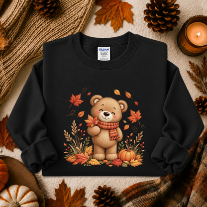A black long-sleeve crewneck sweatshirt with a graphic of a cute cartoon teddy bear surrounded by autumn leaves, pumpkins, and a red scarf. The sweatshirt is presented in a flat-lay arrangement with cozy fall props including pinecones, a lit candle, a knit blanket, and a plaid scarf.