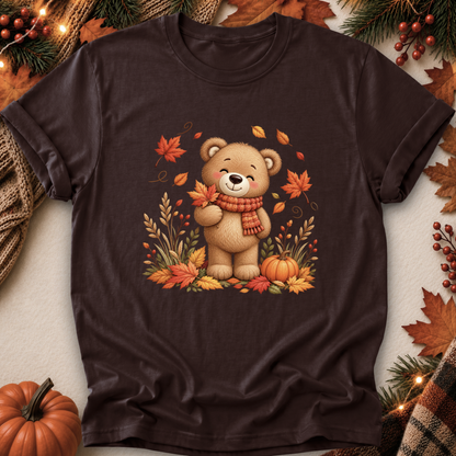 A dark brown short-sleeve t-shirt with a graphic of a cute cartoon teddy bear wearing an orange scarf, surrounded by autumn leaves, pumpkins, and pine branches, presented in a flat-lay setting.