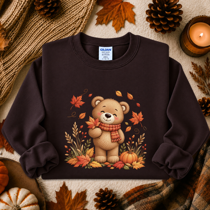 A dark brown crewneck sweatshirt featuring a cute cartoon bear wearing a red scarf and surrounded by autumn leaves and pumpkins, presented in a cozy fall flat-lay setting with pinecones, a candle, and a knit blanket.
