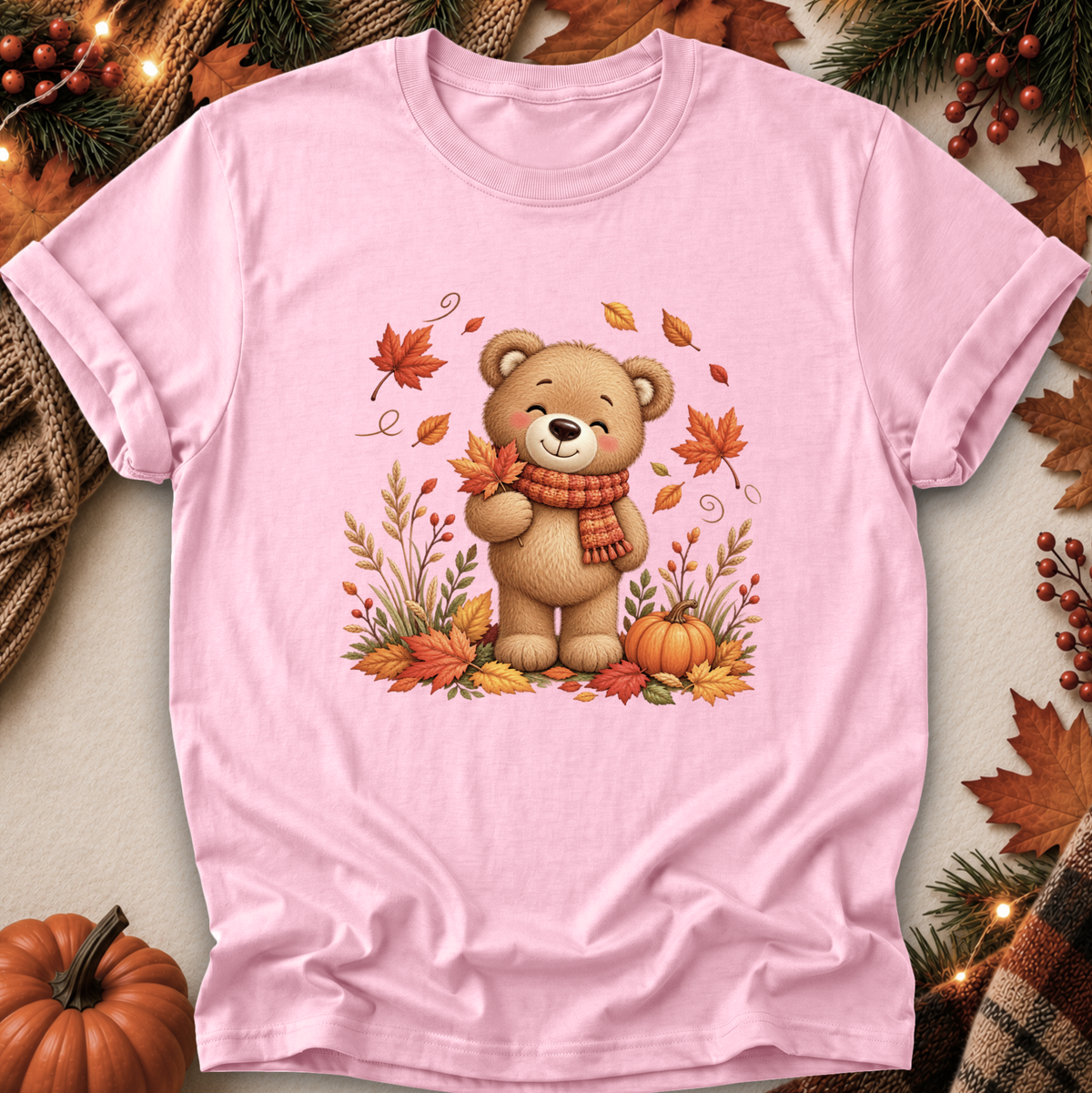 A light pink short-sleeve t-shirt with a central graphic of a cute cartoon bear wearing a red and orange striped scarf, surrounded by autumn leaves, pumpkins, and pine branches. The shirt is displayed in a flat-lay arrangement with seasonal props like a small pumpkin, pinecones, and a plaid scarf.