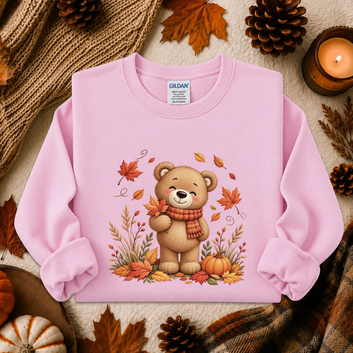 A light pink long-sleeve crewneck sweatshirt laid flat on a cozy autumn-themed background. The sweatshirt features a central graphic of a cute cartoon teddy bear wearing an orange and red striped scarf, surrounded by colorful fall leaves, pinecones, pumpkins, and a knitted blanket.