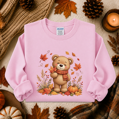 A light pink long-sleeve crewneck sweatshirt laid flat on a cozy autumn-themed background. The sweatshirt features a central graphic of a cute cartoon teddy bear wearing an orange and red striped scarf, surrounded by colorful fall leaves, pinecones, pumpkins, and a knitted blanket.