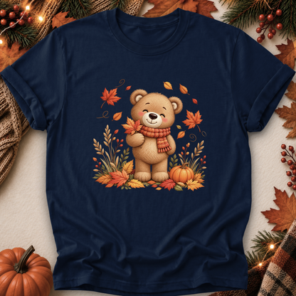 A navy blue short-sleeve t-shirt with a central graphic of a cute cartoon teddy bear wearing a red and orange striped scarf, surrounded by autumn leaves, pumpkins, and pine branches. The shirt is displayed in a flat-lay arrangement with seasonal props like a small pumpkin, pinecones, and string lights on a neutral background.