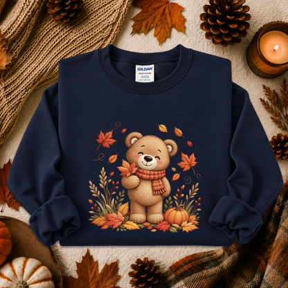 A navy blue crewneck sweatshirt with a graphic of a cute cartoon teddy bear wearing a red scarf, surrounded by autumn leaves, pumpkins, and pinecones. The sweatshirt is presented in a flat-lay arrangement with cozy fall-themed props including a candle, a knit blanket, and a plaid scarf.