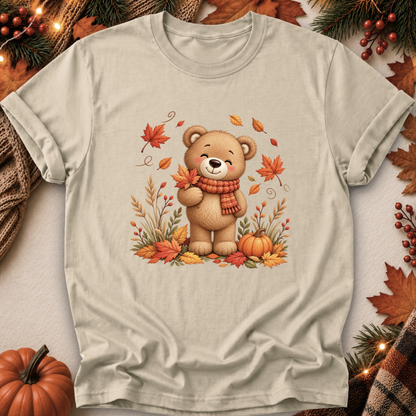 A light beige short-sleeve t-shirt with a central graphic of a smiling brown teddy bear wearing an orange scarf, surrounded by autumn leaves, pumpkins, and pine branches. The shirt is styled in a flat-lay with seasonal props like a small pumpkin, a knit blanket, and fall leaves.