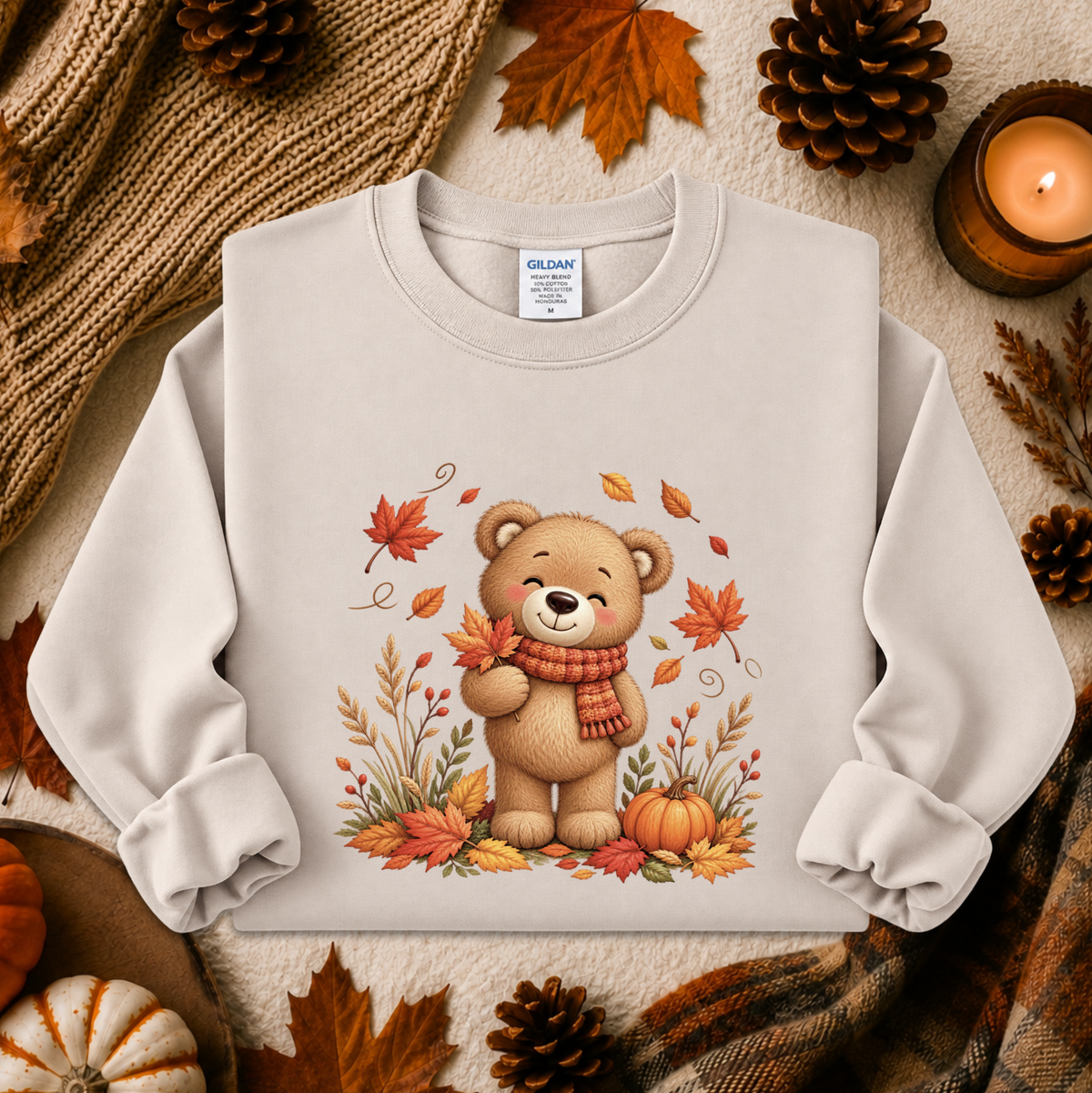 A light beige crewneck sweatshirt featuring a cute cartoon teddy bear wearing a red and orange striped scarf, surrounded by autumn leaves, pumpkins, and pinecones. The sweatshirt is presented in a cozy flat-lay arrangement with a knitted sweater, a lit candle, and fall leaves.