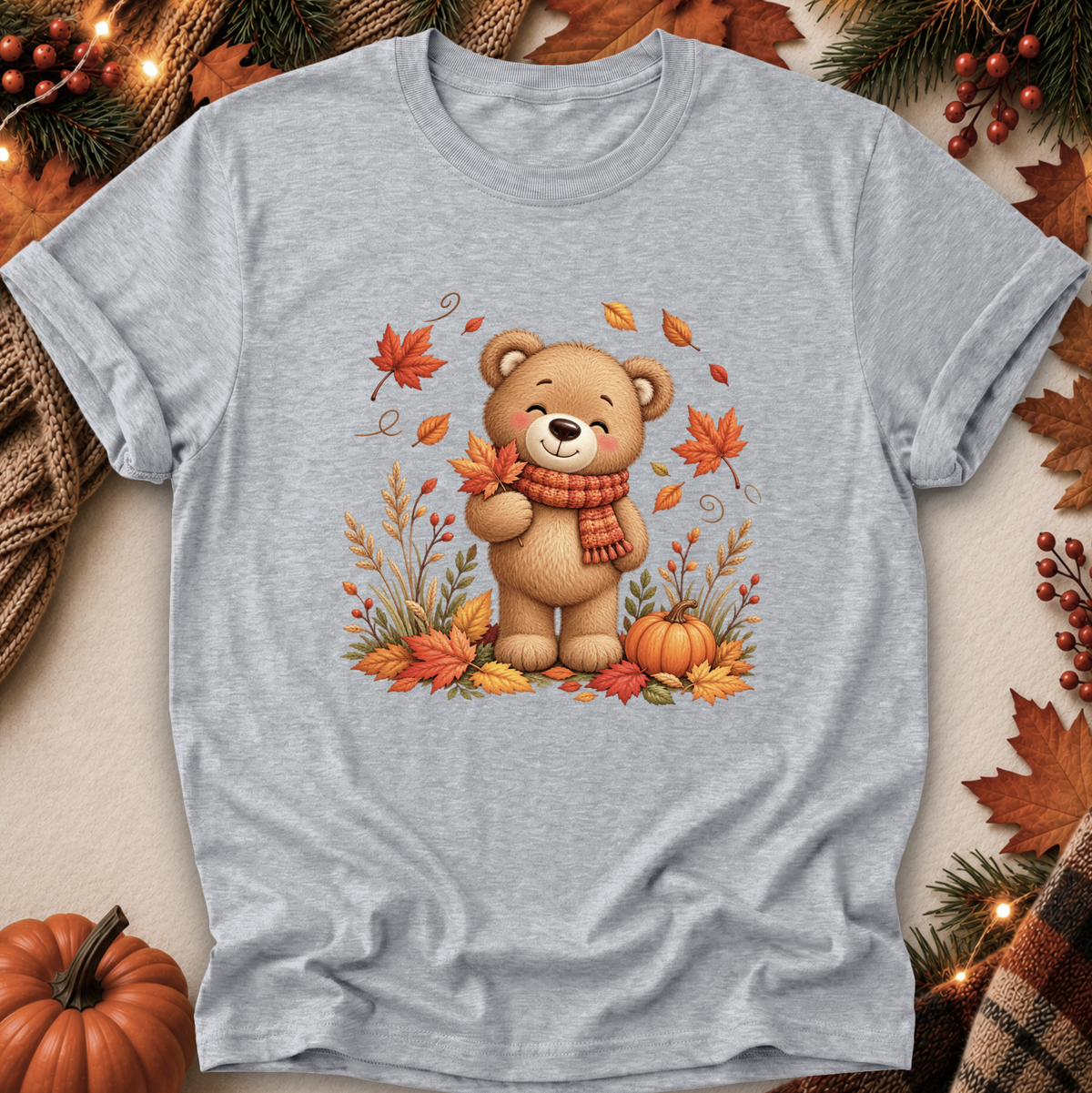 A light heather gray short-sleeve t-shirt with a central graphic of a cute cartoon bear wearing an orange scarf, surrounded by autumn leaves, pumpkins, and fall foliage. The t-shirt is displayed in a flat-lay arrangement with seasonal props like a small pumpkin, pine branches, and a plaid scarf.