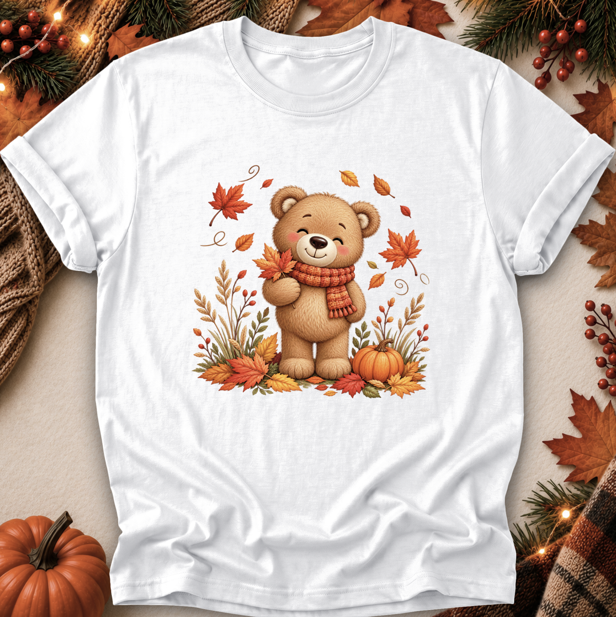 A white short-sleeve crewneck t-shirt featuring a cute cartoon illustration of a smiling brown teddy bear wearing an orange scarf, surrounded by autumn leaves, pumpkins, and grass. The shirt is displayed in a flat-lay setting with seasonal props like a small pumpkin, pine branches, and a plaid scarf.