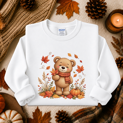 A white long-sleeve crewneck sweatshirt by Gildan, featuring a cute cartoon bear wearing a red and orange striped scarf, surrounded by autumn leaves, pumpkins, and pinecones. The sweatshirt is presented in a flat-lay arrangement with cozy fall-themed props like a knitted blanket, a lit candle, and dried leaves.