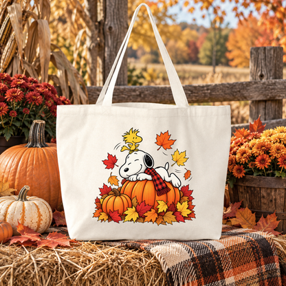 A white canvas tote bag featuring a cartoon illustration of Snoopy and Woodstock on an orange pumpkin surrounded by autumn leaves, displayed in a fall-themed outdoor setting with pumpkins, flowers, and a plaid blanket.