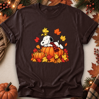 A dark brown short-sleeve t-shirt featuring a graphic of Snoopy resting on a pumpkin surrounded by colorful autumn leaves and pine branches, styled in a flat-lay with seasonal props.