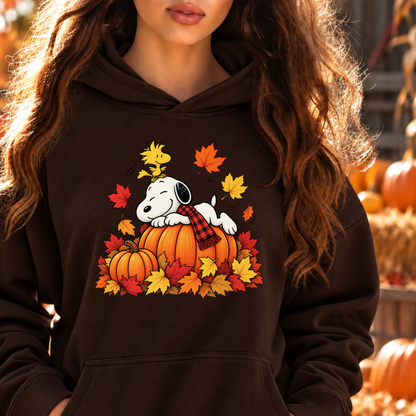 A woman wearing a dark brown hoodie with a colorful graphic of Snoopy and Woodstock on a pumpkin surrounded by autumn leaves. The background is a warm, blurred outdoor fall scene with pumpkins.