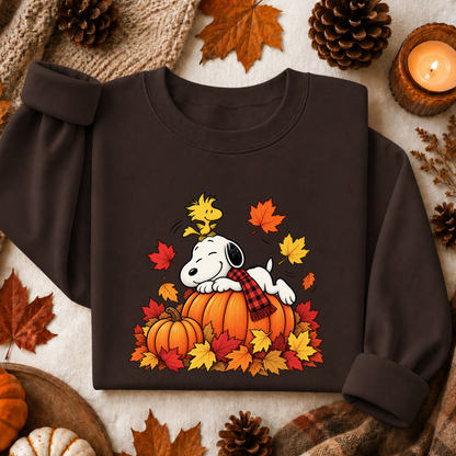 A dark brown crewneck sweatshirt featuring a graphic of Snoopy resting on a pumpkin surrounded by autumn leaves and a plaid scarf, styled in a flat lay with fall-themed props like pinecones, a candle, and leaves.