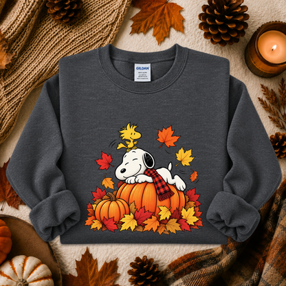 A dark gray crewneck sweatshirt featuring a graphic of Snoopy resting on a pumpkin surrounded by autumn leaves and a plaid scarf, styled in a cozy fall flat-lay with pinecones, a candle, and a knit blanket.