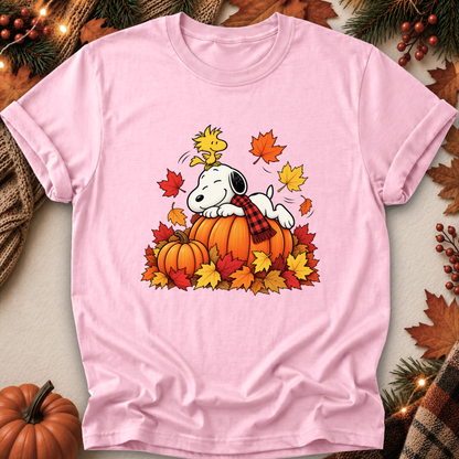 A light pink short-sleeve t-shirt featuring a cartoon graphic of Snoopy resting on an orange pumpkin surrounded by colorful autumn leaves, with Woodstock perched on Snoopy's head. The shirt is displayed in a flat-lay setting with seasonal props like pine branches, a small pumpkin, and a plaid scarf.