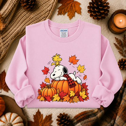 A light pink long-sleeve crewneck sweatshirt featuring a cartoon graphic of Snoopy lying on a pumpkin with Woodstock and autumn leaves. The sweatshirt is styled in a flat lay with fall-themed props including pinecones, a candle, and a knitted blanket.