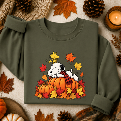 An olive green long-sleeve sweatshirt featuring a graphic of Snoopy resting on a pumpkin with Woodstock and autumn leaves, styled in a flat-lay with fall-themed props like pinecones, leaves, and a candle.