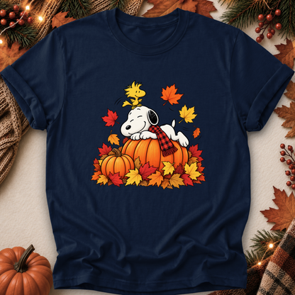 A navy blue short-sleeve t-shirt featuring a graphic of Snoopy resting on a pumpkin surrounded by colorful autumn leaves and a plaid scarf. The shirt is displayed in a flat-lay arrangement with seasonal props including pine branches, a small pumpkin, and lights.