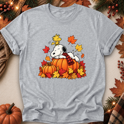 A heather gray short-sleeve t-shirt featuring a cartoon graphic of Snoopy resting on an orange pumpkin surrounded by colorful autumn leaves and a plaid scarf, with Woodstock above. The shirt is displayed in a flat-lay setting decorated with fall leaves, pine branches, and a small pumpkin.