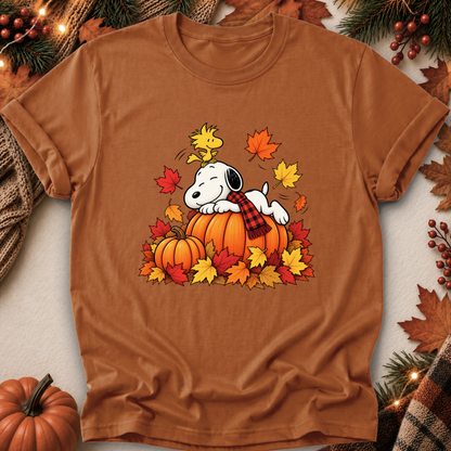 A brown short-sleeve crewneck t-shirt featuring a cartoon graphic of Snoopy resting on an orange pumpkin surrounded by colorful autumn leaves and a plaid scarf. The shirt is displayed in a flat-lay arrangement with seasonal props including a small pumpkin, pine branches, and a plaid knit blanket.
