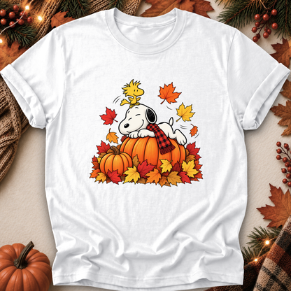 A white short-sleeve crewneck t-shirt featuring a colorful graphic of Snoopy and Woodstock on pumpkins surrounded by autumn leaves. The shirt is displayed in a flat-lay arrangement with seasonal props like a small pumpkin, pine branches, and a plaid scarf.