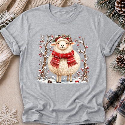 A heather gray short-sleeve t-shirt with a festive Christmas graphic of a fluffy white sheep wearing a red patterned scarf and a green wreath, surrounded by snow-covered branches with red berries. The t-shirt is displayed in a flat-lay arrangement with cozy holiday props including a cream knit sweater, pinecones, and a plaid scarf on a neutral beige background.