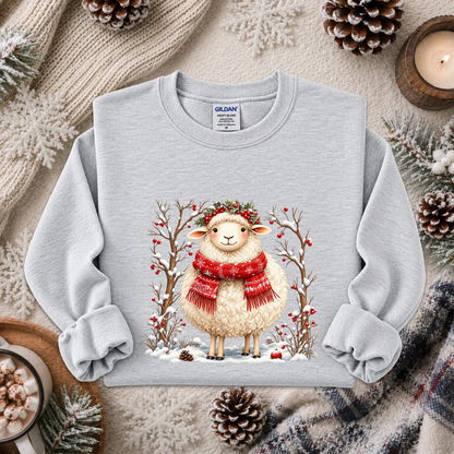 A light gray crewneck sweatshirt featuring a festive graphic of a fluffy white sheep wearing a red knit scarf and a green wreath, surrounded by winter trees with red berries. The sweatshirt is styled in a cozy flat-lay arrangement with a knitted blanket, pinecones, a candle, and a mug of hot cocoa.