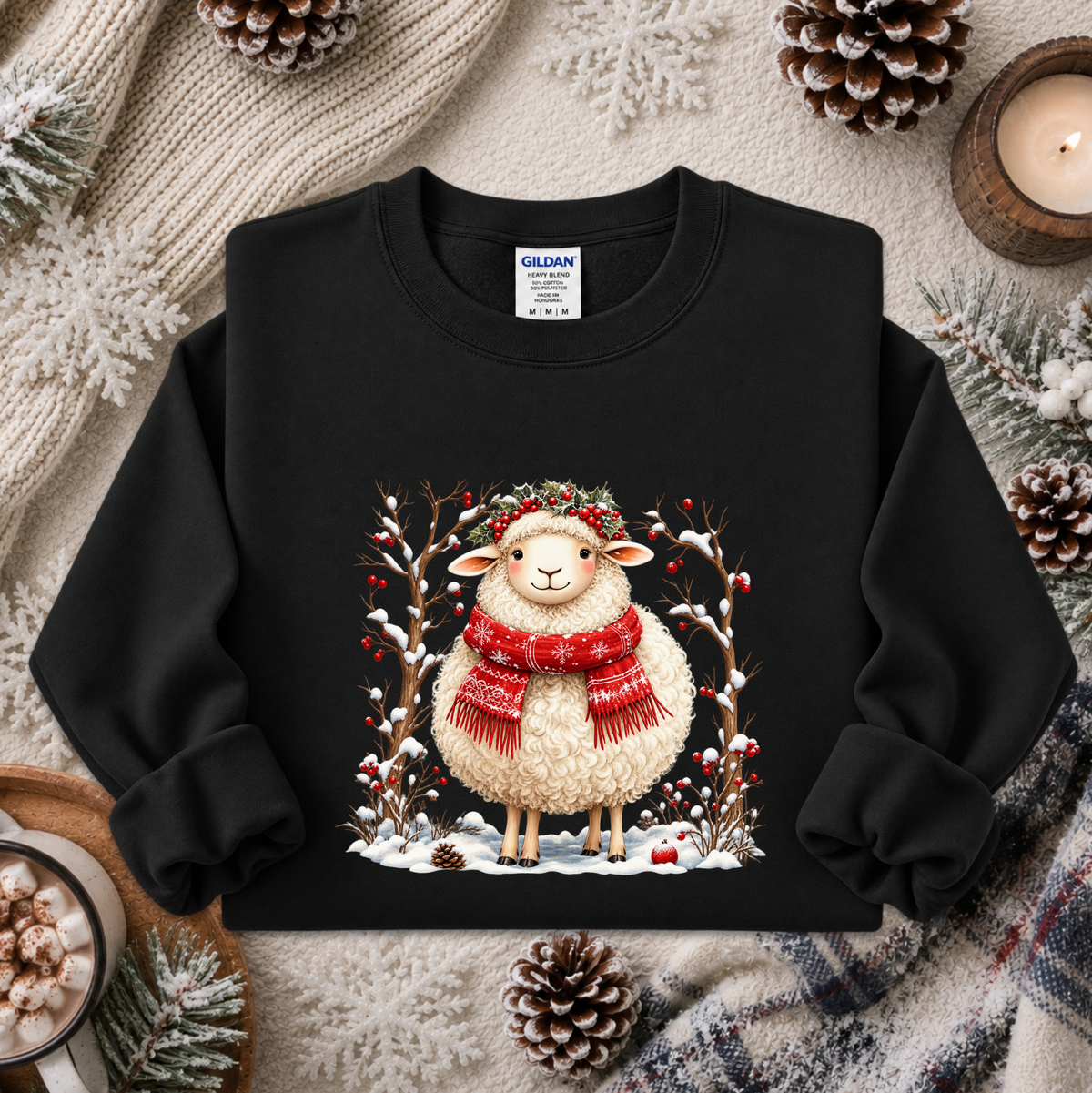 A black crewneck sweatshirt featuring a festive graphic of a fluffy cream-colored sheep wearing a red knit scarf and a green leafy crown, surrounded by snow, berries, and branches. The sweatshirt is styled in a cozy flat-lay arrangement with a knitted blanket, pinecones, a candle, and a mug of hot cocoa.