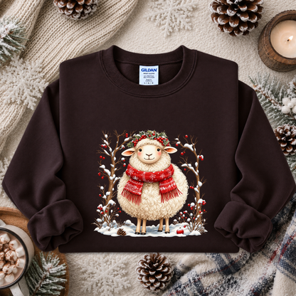 A dark brown crewneck sweatshirt featuring a festive graphic of a fluffy white sheep wearing a red scarf and green wreath, surrounded by snow, pine branches, and red berries. The sweatshirt is styled in a cozy flat-lay arrangement with a knitted beige scarf, pinecones, a lit candle, and a mug of hot cocoa.
