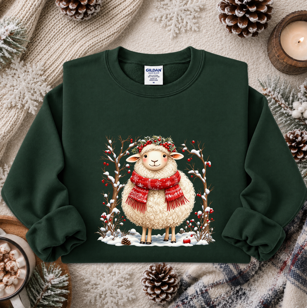 A dark green crewneck sweatshirt featuring a festive graphic of a fluffy white sheep wearing a red knit scarf and a green leafy crown, surrounded by snow, pine branches, and red berries. The sweatshirt is styled in a cozy flat-lay arrangement with a cream knit blanket, pinecones, a lit candle, and a cup of hot cocoa.