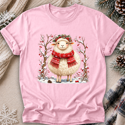 A light pink short-sleeve crewneck t-shirt featuring a festive graphic of a cute white sheep wearing a red snowflake scarf and a green wreath, standing in a snowy winter scene. The t-shirt is displayed in a flat-lay setting with cozy holiday props like pinecones, a knit sweater, and a plaid scarf.