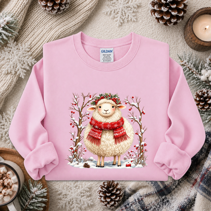 A pink long-sleeve sweatshirt with a graphic of a smiling sheep in a red knit scarf and green leafy crown, surrounded by winter branches and snow. The sweatshirt is displayed in a cozy flat-lay setting with pinecones, a candle, and knitted blankets.