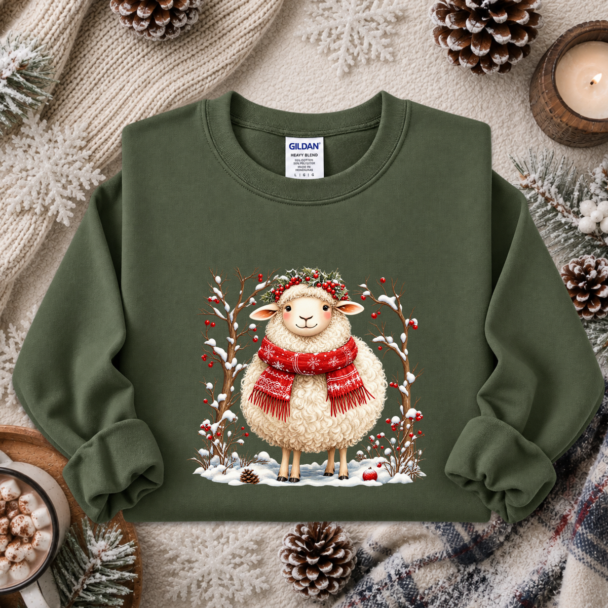 An olive green crewneck sweatshirt featuring a festive graphic of a fluffy white sheep wearing a red knit scarf and a green wreath, surrounded by snow, berries, and pinecones. The sweatshirt is styled in a cozy flat-lay arrangement with a knitted sweater, pinecones, a candle, and a mug of cocoa.