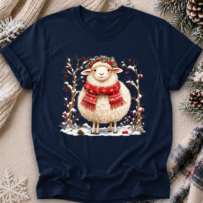 A navy blue short-sleeve t-shirt with a festive Christmas graphic of a fluffy white sheep wearing a red scarf and green wreath, surrounded by snow-covered branches and red berries. The t-shirt is displayed in a flat-lay setting on a cozy beige knit blanket with pinecones and evergreen branches around it.