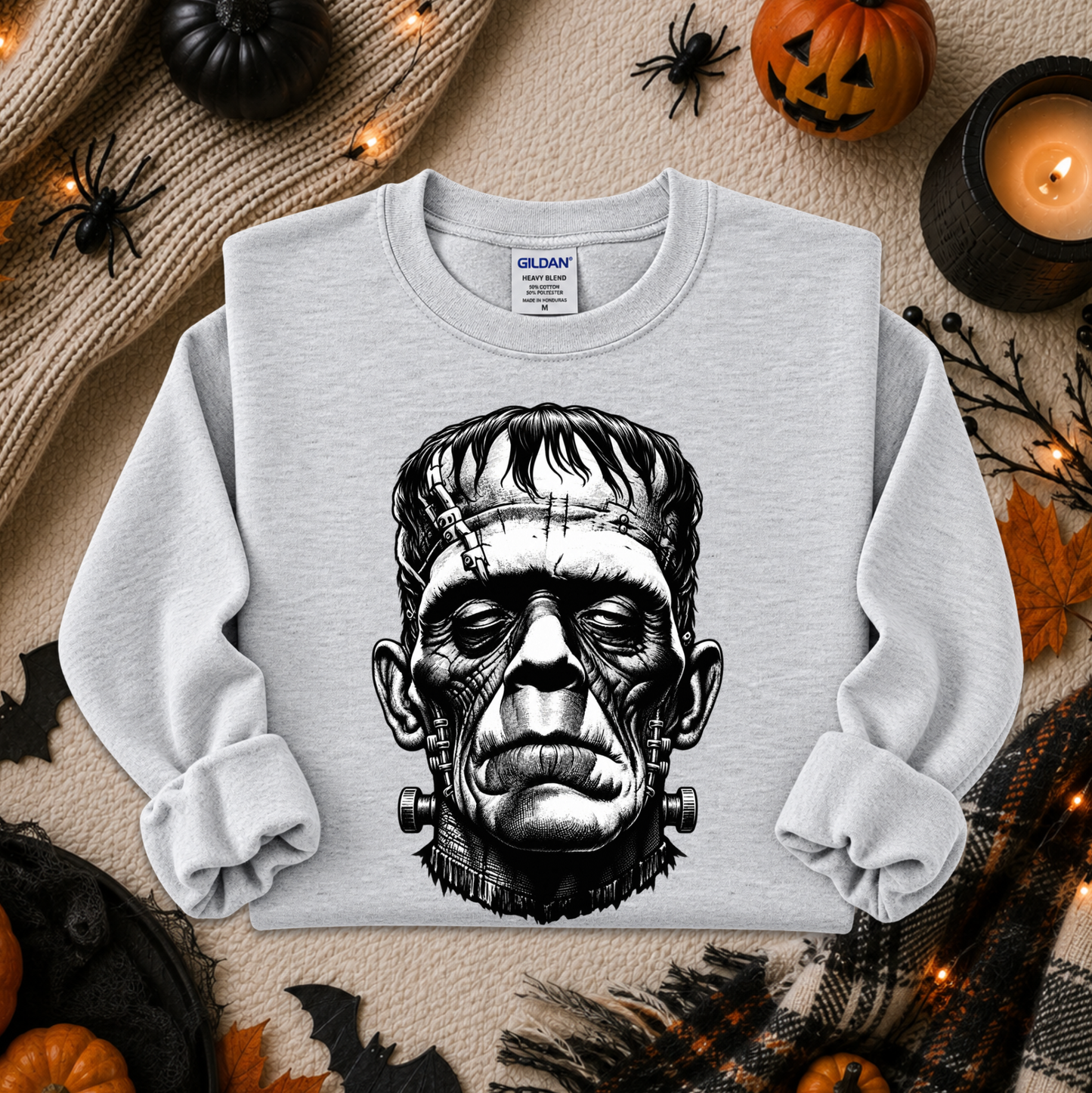 A light gray crewneck sweatshirt with a black and white graphic of Frankenstein's monster, laid flat on a cozy beige blanket surrounded by Halloween decorations including pumpkins, spiderwebs, and a candle.